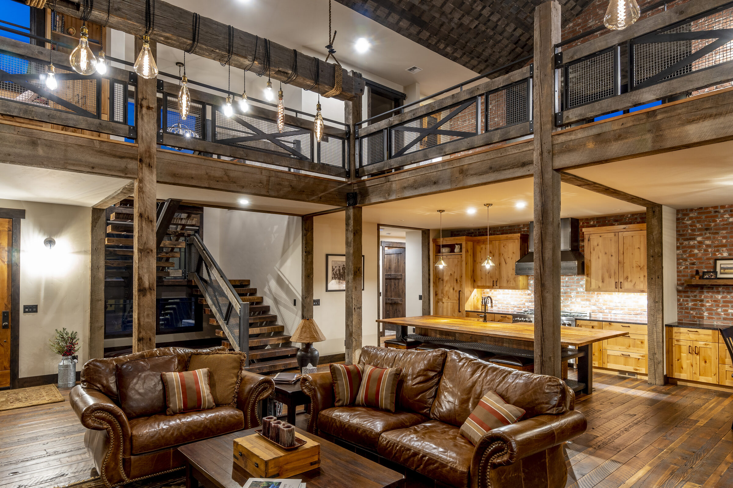 Two-story great room framed by heavy timber posts, a barn-wood open stair, and a mesh-railed loft, with a knotty-alder-and-brick kitchen beyond