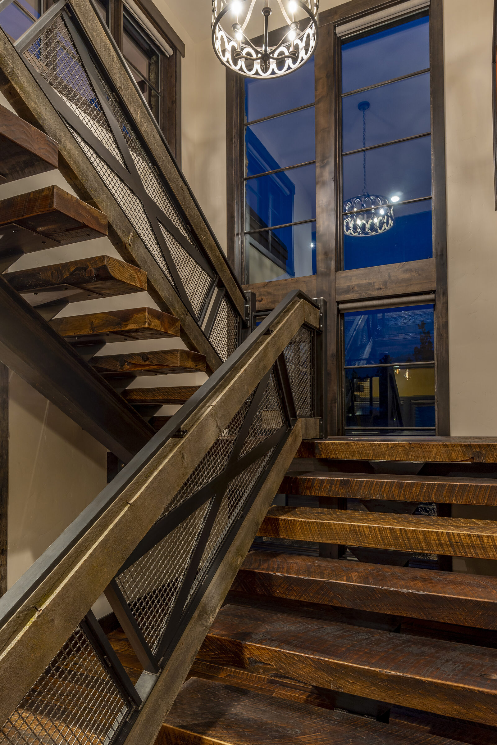 Open-tread staircase milled from 130-year-old barn wood with blackened-steel railings, beneath a wrought-iron chandelier