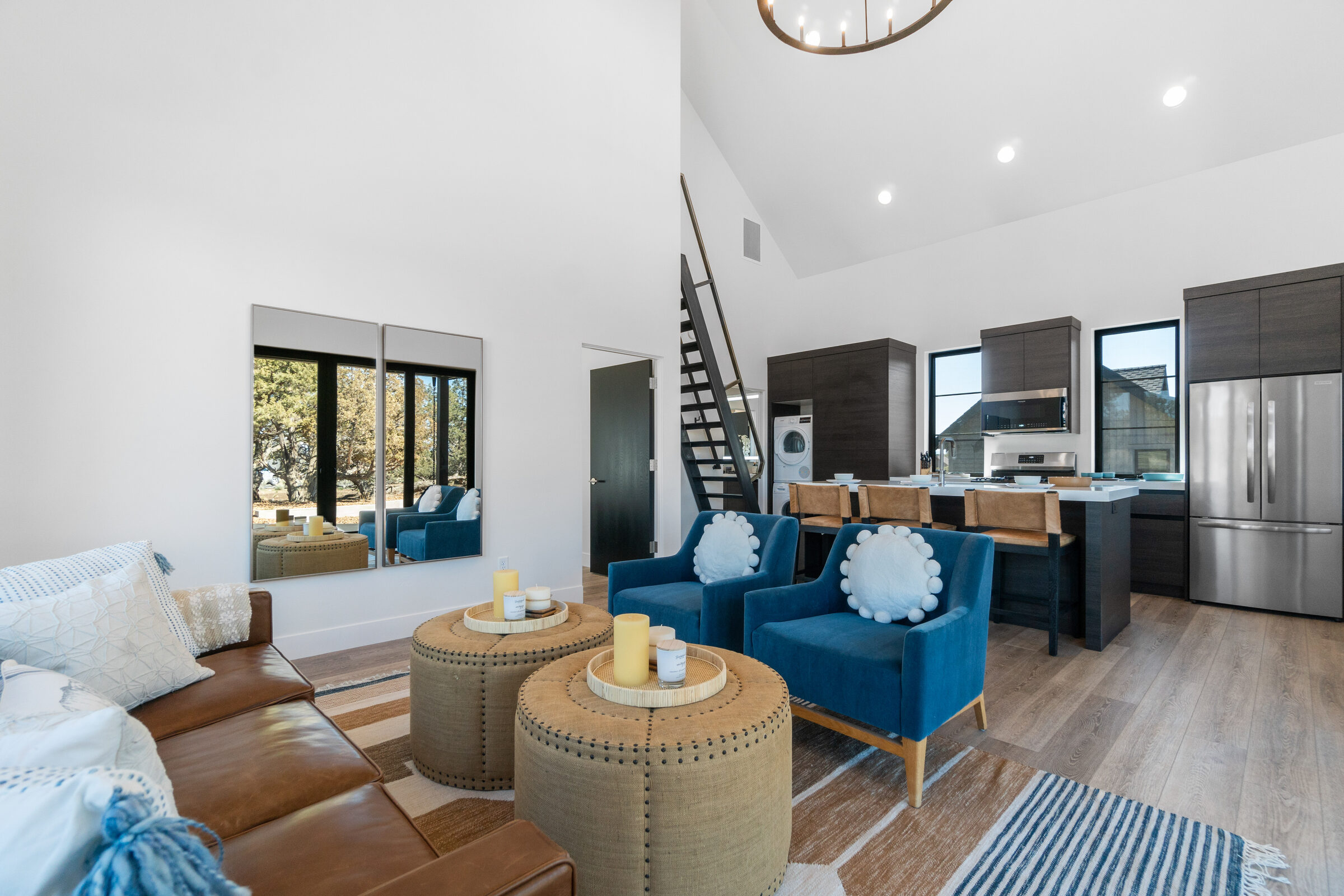 Living room with a leather sectional and blue lounge chairs, open to the kitchen and stair beneath walls of glass