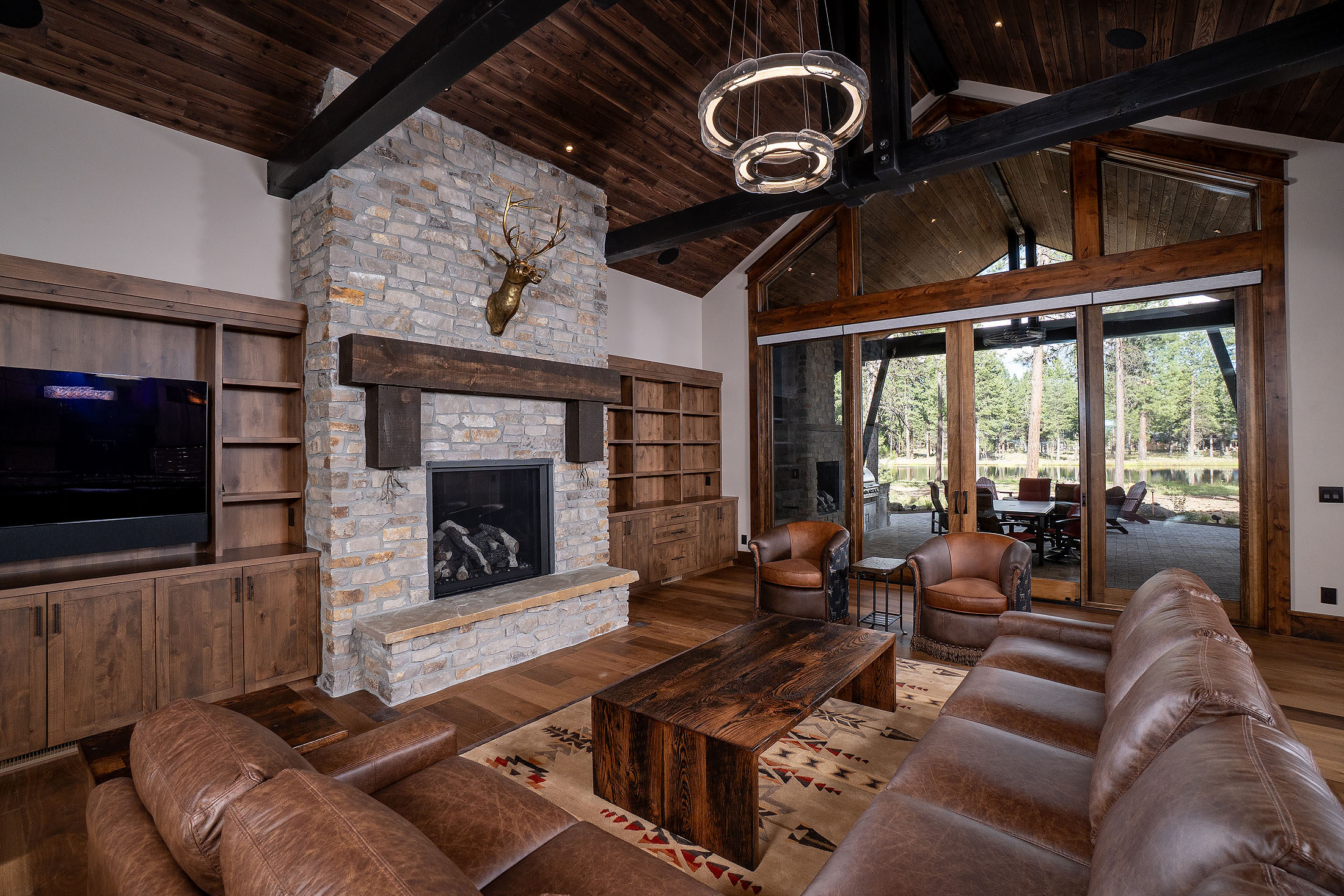 Great room with a stacked-stone fireplace, timber beams, and leather seating open to the forest